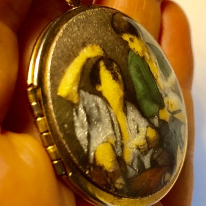 May include: Gold-tone oval locket pendant with a detailed miniature painting inside. The painting depicts figures in shades of yellow, green, and gray. The locket is suspended from a delicate gold chain, creating a classic jewelry piece.
