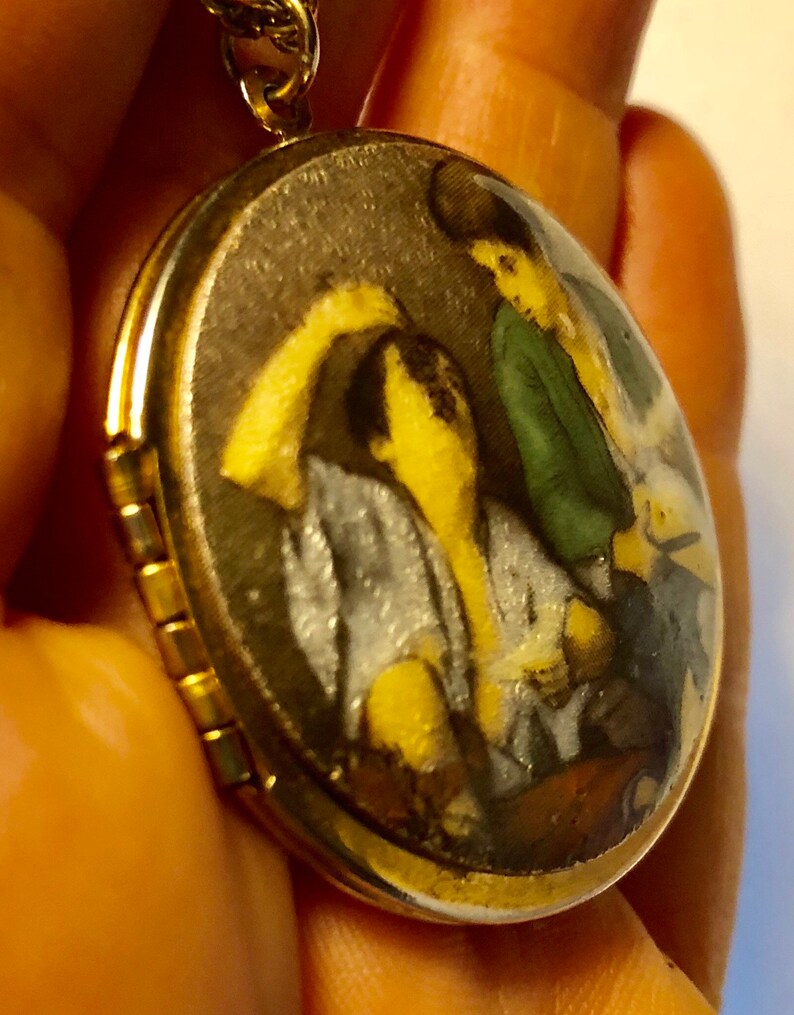 May include: Gold-tone oval locket pendant with a detailed miniature painting inside. The painting depicts figures in shades of yellow, green, and gray. The locket is suspended from a delicate gold chain, creating a classic jewelry piece.