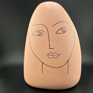 以下が含まれることがあります： 黒い線画の顔を描いた薄いピンクの陶器の彫刻。彫刻は卵の形をしている。