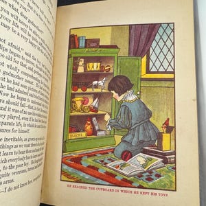 May include: A vintage book illustration depicts a child kneeling before a green cupboard full of toys. The text below reads, "HE REACHED THE CUPBOARD IN WHICH HE KEPT HIS TOYS." The scene is set in a room with a patterned rug.