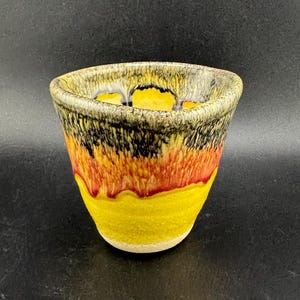 Puede incluir: Una taza de cerámica hecha a mano con una combinación de colores vibrantes. La taza presenta una base amarilla, que pasa al rojo y al naranja, y está rematada con detalles en negro y crema. El interior es amarillo. La taza tiene una forma ligeramente acampanada.