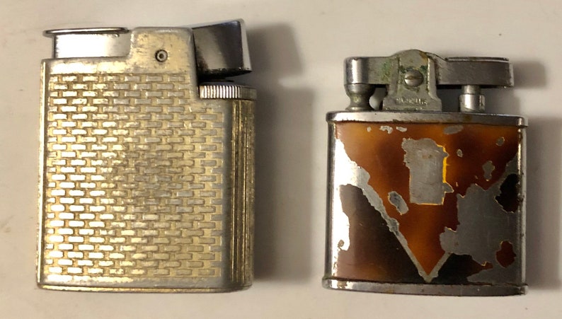 May include: Two vintage lighters, one with a silver finish and a brick pattern, the other with a brown and silver finish with a geometric design.