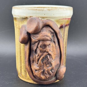 May include: A handcrafted ceramic mug with a tan and cream color scheme. The mug features a detailed relief of a bearded man. The mug has a cylindrical shape with vertical ridges and a cream-colored rim.
