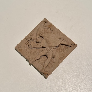 Puede incluir: Un azulejo de cerámica marrón con una escultura en relieve de un bufón que sostiene una flor. El bufón lleva un sombrero puntiagudo y una túnica larga. El azulejo tiene un borde decorativo.