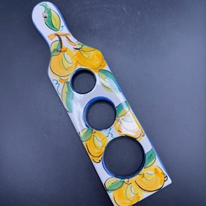May include: A white ceramic serving board with three circular cutouts. The board is decorated with yellow lemons and green leaves, outlined in black, and has a blue border. The handle has a hole for hanging.