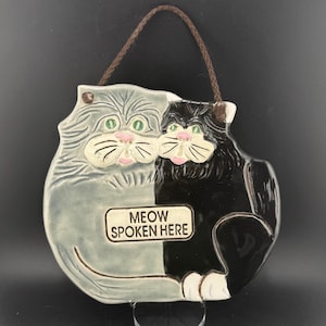 Puede incluir: Adorno de pared de cerámica con forma de gato, mitad gris y mitad negro, con ojos verdes y narices rosas. El texto "MEOW SPOKEN HERE" está en una placa rectangular. Cordón marrón para colgar.