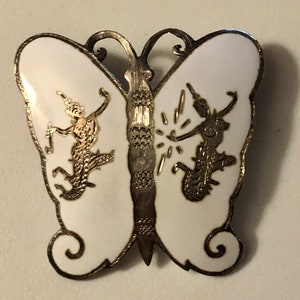 May include: A silver butterfly brooch with white enamel and gold accents. The butterfly has two figures on its wings, each holding a staff.