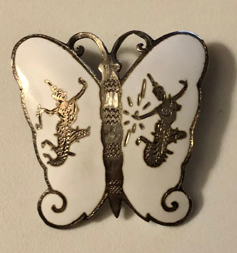 May include: A silver butterfly brooch with white enamel and gold accents. The butterfly has two figures on its wings, each holding a staff.