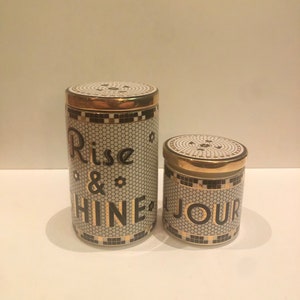 May include: Two ceramic canisters with gold lids and black and white mosaic designs. The larger canister says "Rise & Shine" and the smaller canister says "Jour".