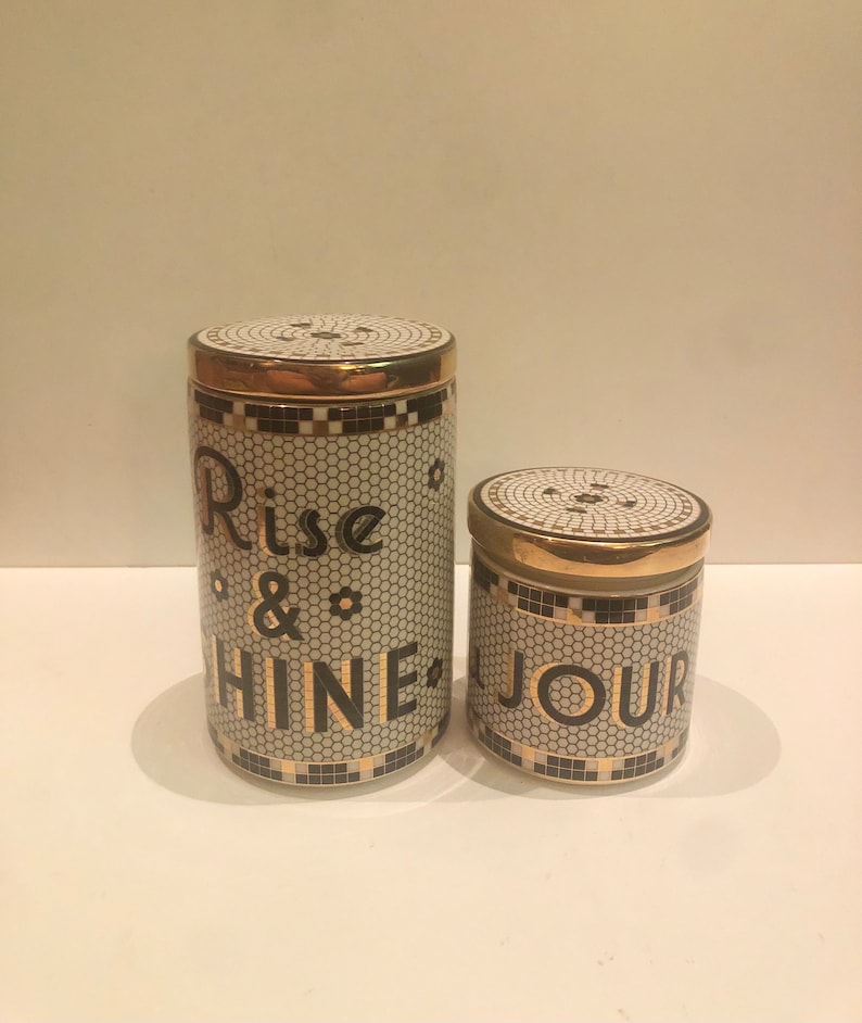 May include: Two ceramic canisters with gold lids and black and white mosaic designs. The larger canister says "Rise & Shine" and the smaller canister says "Jour".