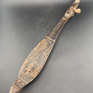 May include: A dark brown, wooden stringed instrument with a long neck and carved body. The instrument features intricate carvings and a tuning peg. The body has a boat-like shape. The instrument appears to be handmade.