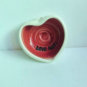 Peut inclure: Bol en céramique en forme de cœur avec un bord blanc et un intérieur rouge. L'intérieur présente un motif en spirale et les mots "LOVE a lot..." en noir. Le bol est posé sur une surface blanche.