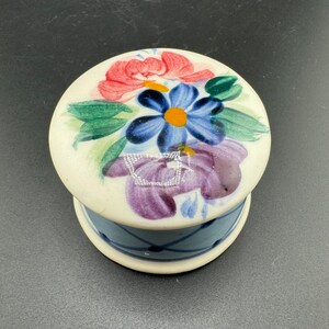 Pode incluir: Uma pequena caixa redonda de cerâmica com um design floral. A tampa é branca com flores pintadas à mão em tons de rosa, azul, verde e roxo. A base da caixa é azul com um padrão de treliça branca.