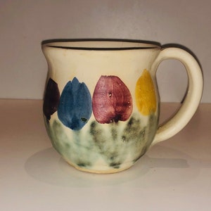 May include: A small, off-white ceramic mug with a rounded body and a handle. The mug is decorated with four tulip designs in shades of purple, blue, and yellow, set against a green and gray base. The mug has a simple, handmade aesthetic.