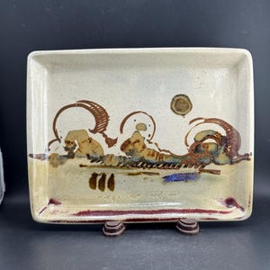 May include: A rectangular ceramic tray with a speckled off-white glaze. The surface features abstract brown and blue painted designs, including circular motifs and horizontal lines. The edges are accented with a dark reddish-brown glaze.