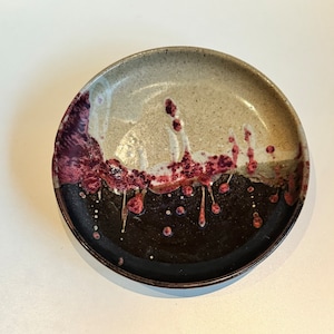 Puede incluir: Un plato de cerámica con un esmalte marrón oscuro y un diseño de goteo blanco y burdeos.