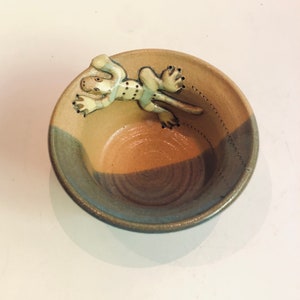 May include: A small, brown and blue ceramic bowl with a white glazed lizard design on the rim. The bowl has a speckled glaze and a brown interior.