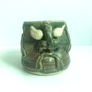 May include: A green ceramic mug with a face sculpted on the front. The face has a white mustache, eyebrows, and nose. The mug has a brown glaze.