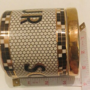 May include: A white ceramic sugar jar with a gold lid and black and gold geometric pattern. The jar has the word "Sugar" written on it in black and gold letters.