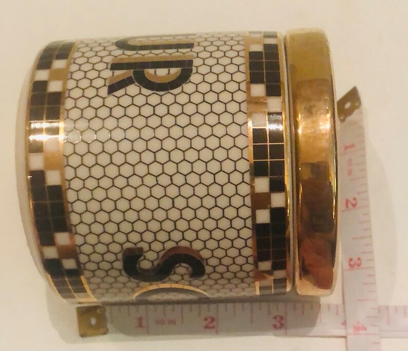 May include: A white ceramic sugar jar with a gold lid and black and gold geometric pattern. The jar has the word "Sugar" written on it in black and gold letters.