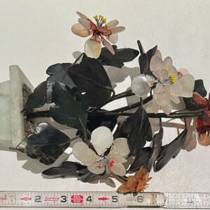 May include: A decorative floral sculpture featuring white, pink, and brown flowers with yellow centers, set against dark green leaves. The sculpture is mounted on a light green rectangular base. A measuring tape shows the sculpture is approximately 6 inches tall.