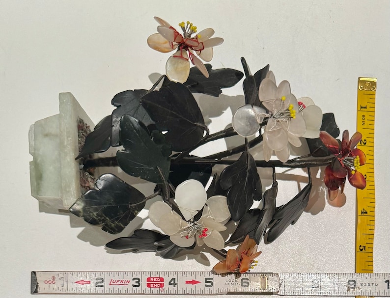 May include: A decorative floral sculpture featuring white, pink, and brown flowers with yellow centers, set against dark green leaves. The sculpture is mounted on a light green rectangular base. A measuring tape shows the sculpture is approximately 6 inches tall.