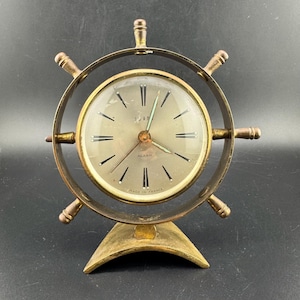 May include: Vintage brass alarm clock in the shape of a ship's wheel. The clock face is gold with black numerals and hands. The words "ALARM" and "MADE IN FRANCE" are visible on the clock face. The clock has a brass stand.