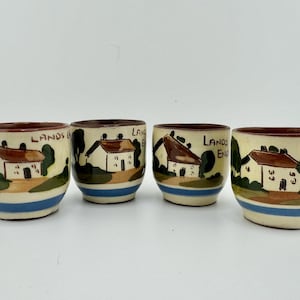 May include: Set of four ceramic egg cups with a white background and brown, green, and blue painted details. Each cup features a cottage with a red roof and the text "Lands End" on the side.
