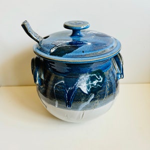 May include: A blue and white ceramic crock with a lid and a small spoon attached to the lid. The crock has a white base and blue and white glaze with a drip effect.