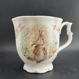 以下が含まれることがあります： クリーム色の陶器のティーカップ。茶色のジャケットを着たウサギが葉の中にいる詳細なイラストが描かれています。装飾的なハンドルとスカラップベースがあり、ティータイムに最適です。