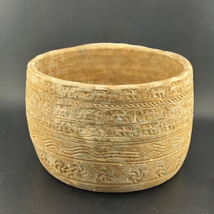 May include: A beige, cylindrical ceramic pot with detailed, carved horizontal bands. The bands feature repeating patterns of stylized floral and wave designs. The pot has a wide opening and a textured surface, suitable for plants or decorative use.