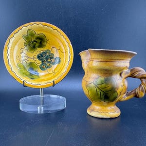 May include: A set of two ceramic pieces with a grapevine design. The larger piece is a pitcher with a twisted handle. The smaller piece is a plate with a scalloped edge.
