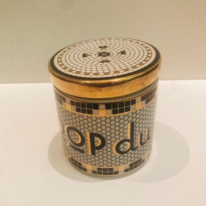 May include: A white ceramic jar with a gold lid and black and gold mosaic design. The jar has the words "pop du" written in gold letters on the front.