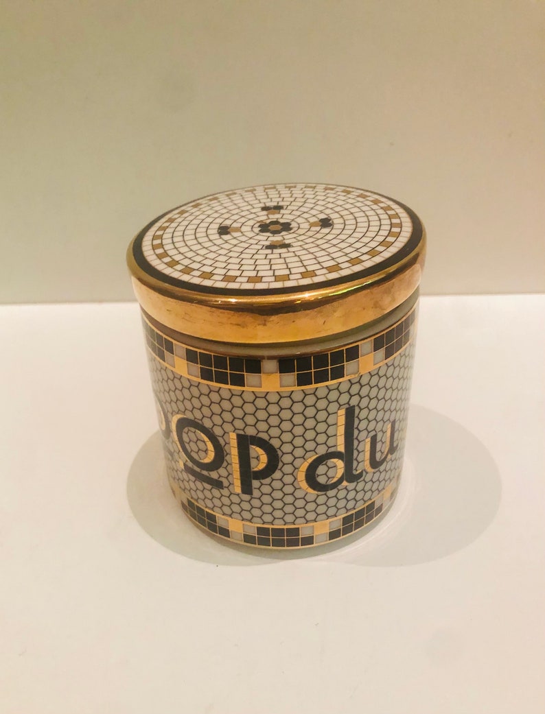 May include: A white ceramic jar with a gold lid and black and gold mosaic design. The jar has the words "pop du" written in gold letters on the front.