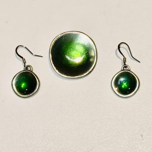 Puede incluir: Un conjunto de pendientes y un broche de color verde y plateado. Los pendientes y el broche tienen forma redonda con un centro de esmalte verde y un borde plateado.
