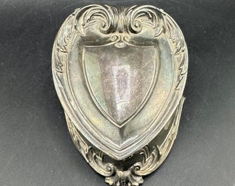 May include: Silver-toned, heart-shaped decorative dish with ornate scrollwork and a central shield design. The dish has a detailed, vintage aesthetic, perfect for holding small items or as a decorative accent. The dish is made of metal.