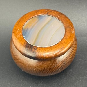 May include: A small, round wooden box with a lid. The lid has a circular inset of polished stone with bands of color. The wood grain is visible, and the box is a warm brown color.