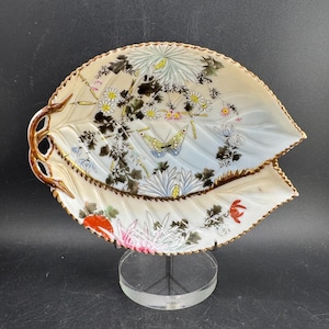 May include: A pair of leaf-shaped decorative porcelain dishes. The dishes are white with hand-painted floral and butterfly designs in shades of yellow, pink, and blue. The edges are trimmed with a gold-colored border. The dishes are displayed on a clear stand.