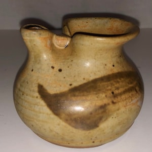 May include: A small, hand-thrown ceramic pitcher with a brown glaze and a speckled finish. The pitcher has a unique, asymmetrical spout and a rounded body.