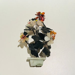 Puede incluir: Un arreglo floral decorativo en una maceta cuadrada de color verde claro. Las flores son blancas, naranjas y rojas con centros amarillos. Las hojas son de color verde oscuro. La maceta y las flores parecen ser de piedra.