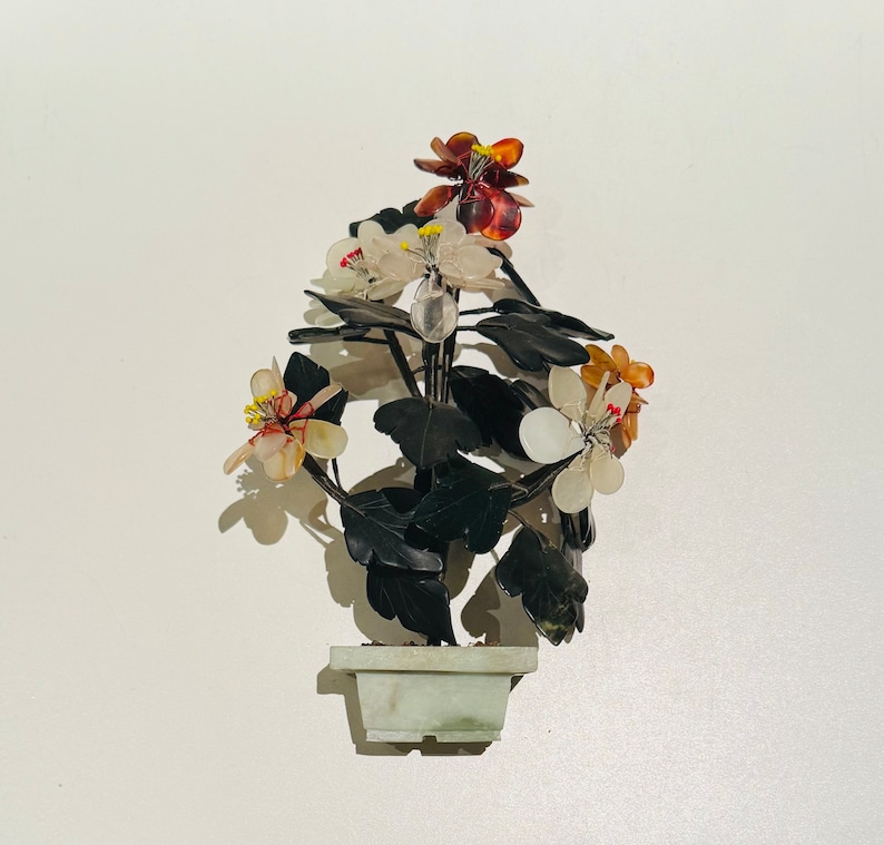 May include: A decorative floral arrangement in a square, light green pot. The flowers are white, orange, and red with yellow centers. The leaves are dark green. The pot and flowers appear to be made of stone.