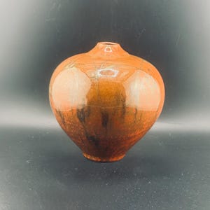 May include: A round, ceramic vase with a glossy, reddish-brown finish. The vase has a narrow neck and a bulbous body. The color transitions from a lighter orange at the top to a darker brown at the base, with some black accents.