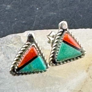May include: A pair of triangular stud earrings. Each earring features a silver-toned rope border, and a design of red and turquoise-colored stones. The earrings are set against a light-colored surface.