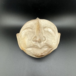 Puede incluir: Una máscara de madera blanca con un rostro tallado, ojos cerrados y una amplia sonrisa. La máscara está hecha de madera clara y tiene un acabado liso y pulido.