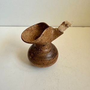 Pode incluir: Um pequeno vaso de cerâmica marrom com um bico curvo único e um cordão branco preso ao topo. O vaso tem uma superfície texturizada salpicada.