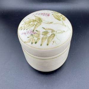 May include: A round, off-white ceramic container with a lid. The lid is decorated with a floral design in shades of green and pink. The container has a cylindrical base and a rounded top, suitable for storing small items.