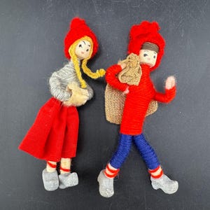 Könnte beinhalten: Zwei handgefertigte Puppen. Eine Puppe trägt einen roten Hut, ein graues Hemd, einen roten Rock und graue Schuhe. Die andere Puppe trägt einen roten Hut, ein rotes Hemd, eine blaue Hose und graue Schuhe und trägt einen Jutesack. Auf schwarzem Hintergrund.