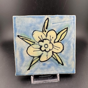 Pode incluir: Azulejo de cerâmica quadrado com fundo azul claro e desenho floral pintado à mão. A flor é creme com contornos pretos e folhas verdes. O azulejo é exibido em um suporte de plástico transparente.