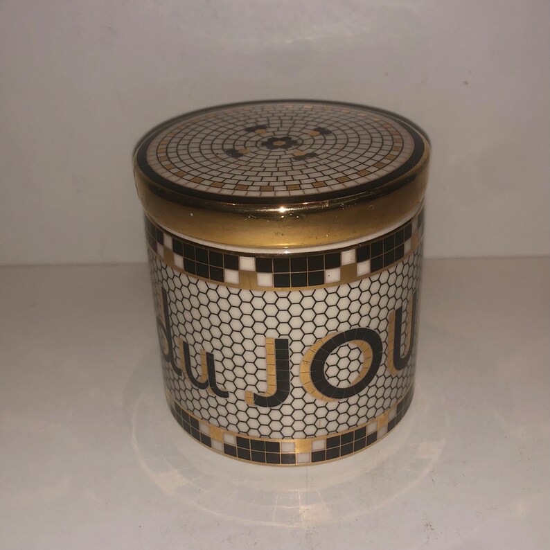 May include: A white ceramic jar with a black and gold mosaic design. The jar has the word "Du Jour" written in gold letters on the side.