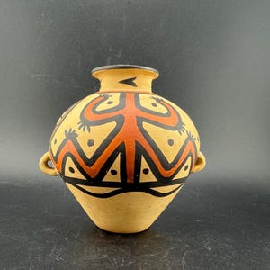 Könnte beinhalten: Eine beige Keramikvase mit rundem Körper und schmalem Hals. Die Vase ist mit einem geometrischen Design in Schwarz und Orange verziert, das eine zentrale Figur und symmetrische Muster aufweist. Zwei kleine Griffe befinden sich an den Seiten.